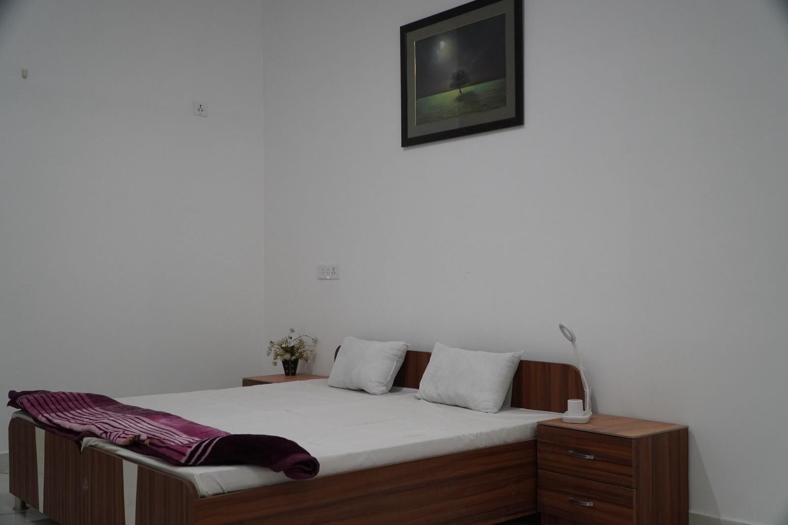 A bedroom with a wooden bed, white bedding, a nightstand with a lamp, and a framed picture on the wall.