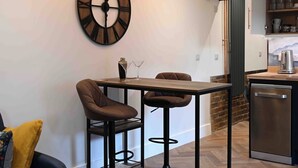 A modern kitchen with a wooden table, two bar stools, a large wall clock, and a dishwasher.