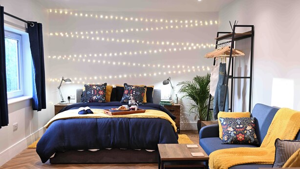 A bedroom with a bed, a sofa, a nightstand, a lamp, a plant, and a wall with string lights.
