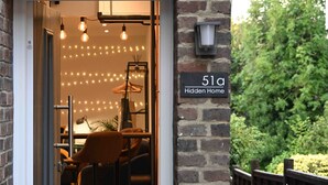 A cozy entrance with a brick wall, a sign reading "51a Hidden Home," and a well-lit interior with hanging lights and furniture.