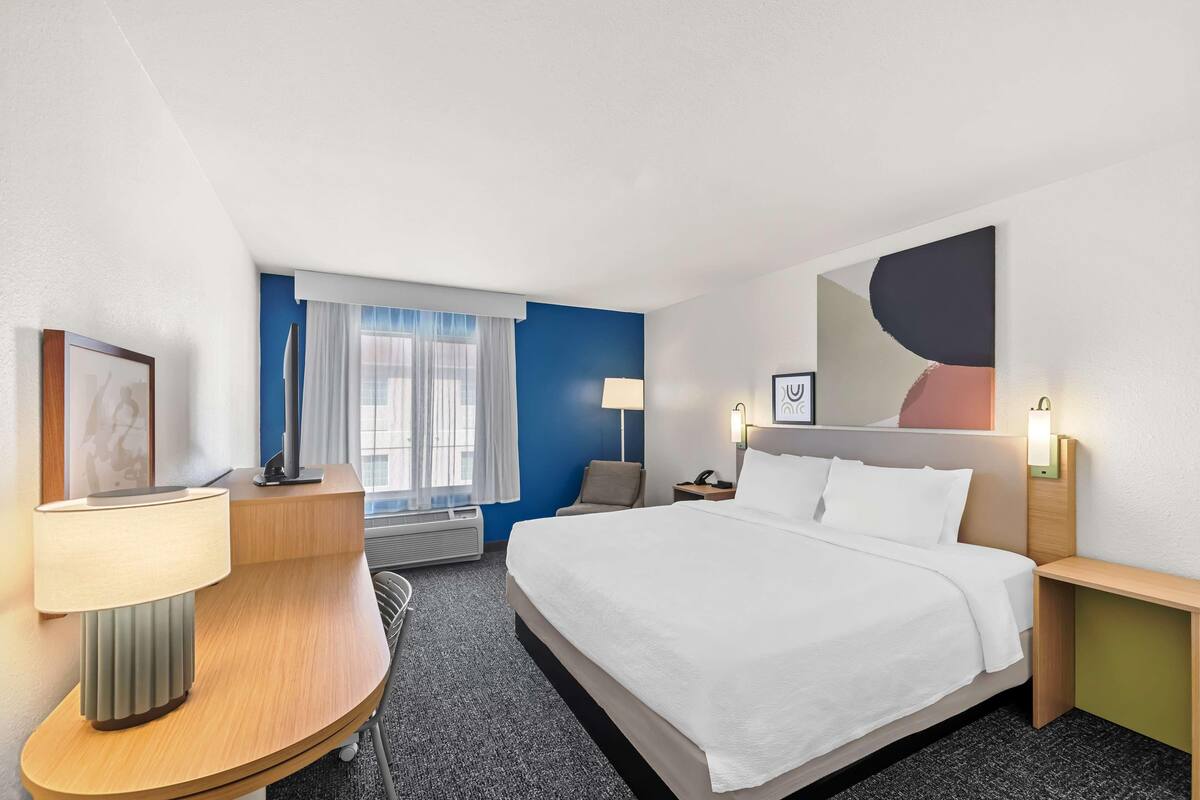 Habitación de hotel con cama, mesitas de noche, escritorio, lámpara, silla, ventana con cortinas y una obra de arte colgada en la pared.
