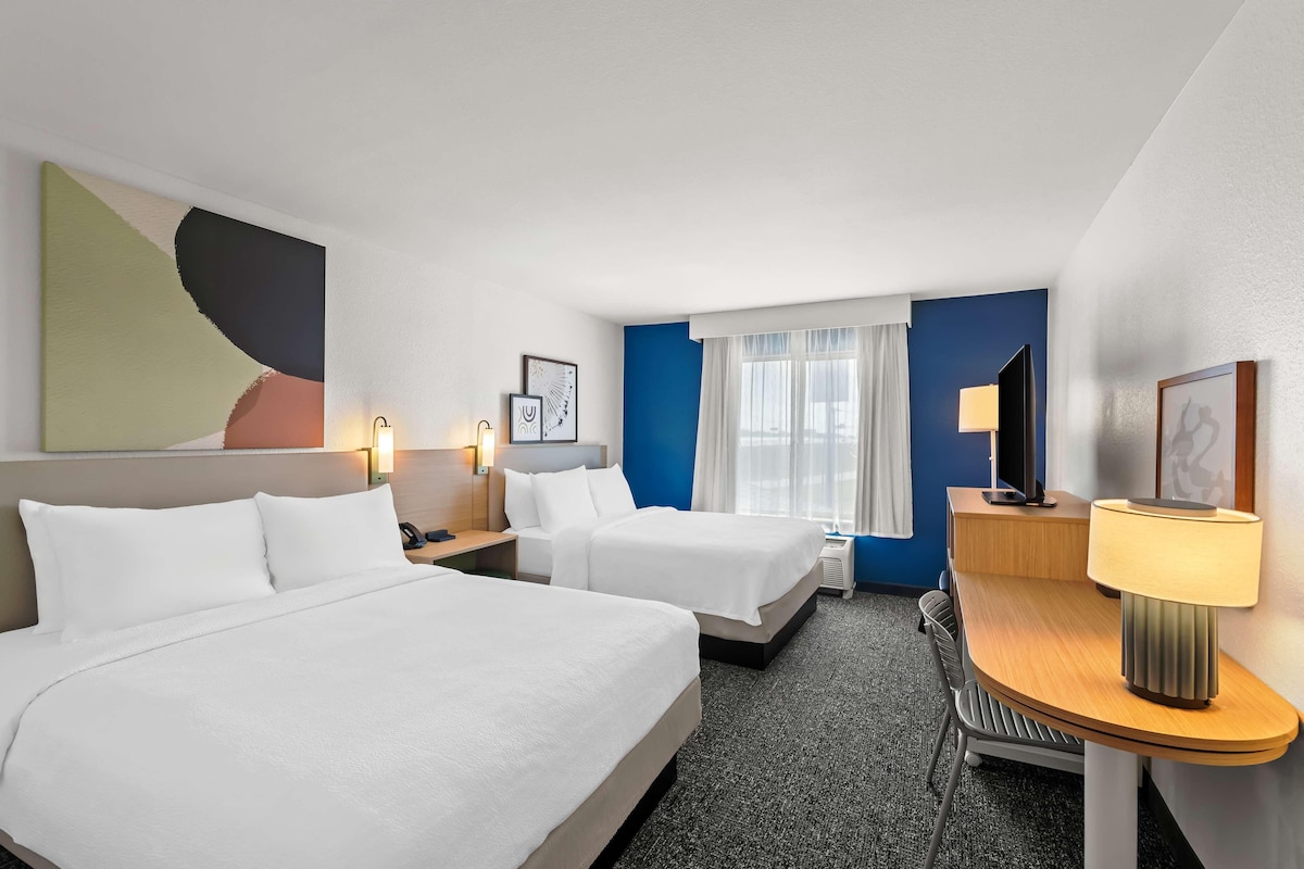 Habitació d'hotel amb dos llits, un escriptori, una cadira, una làmpada i un quadre abstracte gran a la paret.