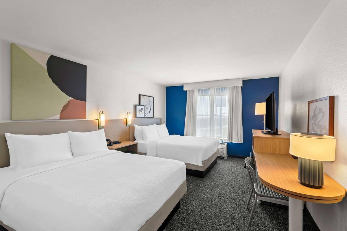 Habitación de hotel con dos camas, un escritorio, una silla, una lámpara y un cuadro abstracto grande en la pared.