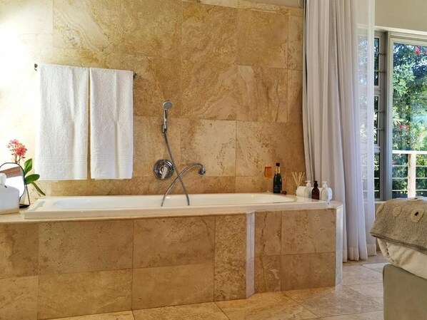 A bathroom with a large bathtub, marble walls, a towel rack, and a window with curtains.