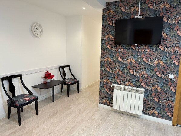 Una sala de estar moderna con un televisor de pantalla plana montado en una pared cubierta de papel tapiz floral, dos sillas de madera oscura, una mesita con un jarrón de flores rojas y un radiador blanco.