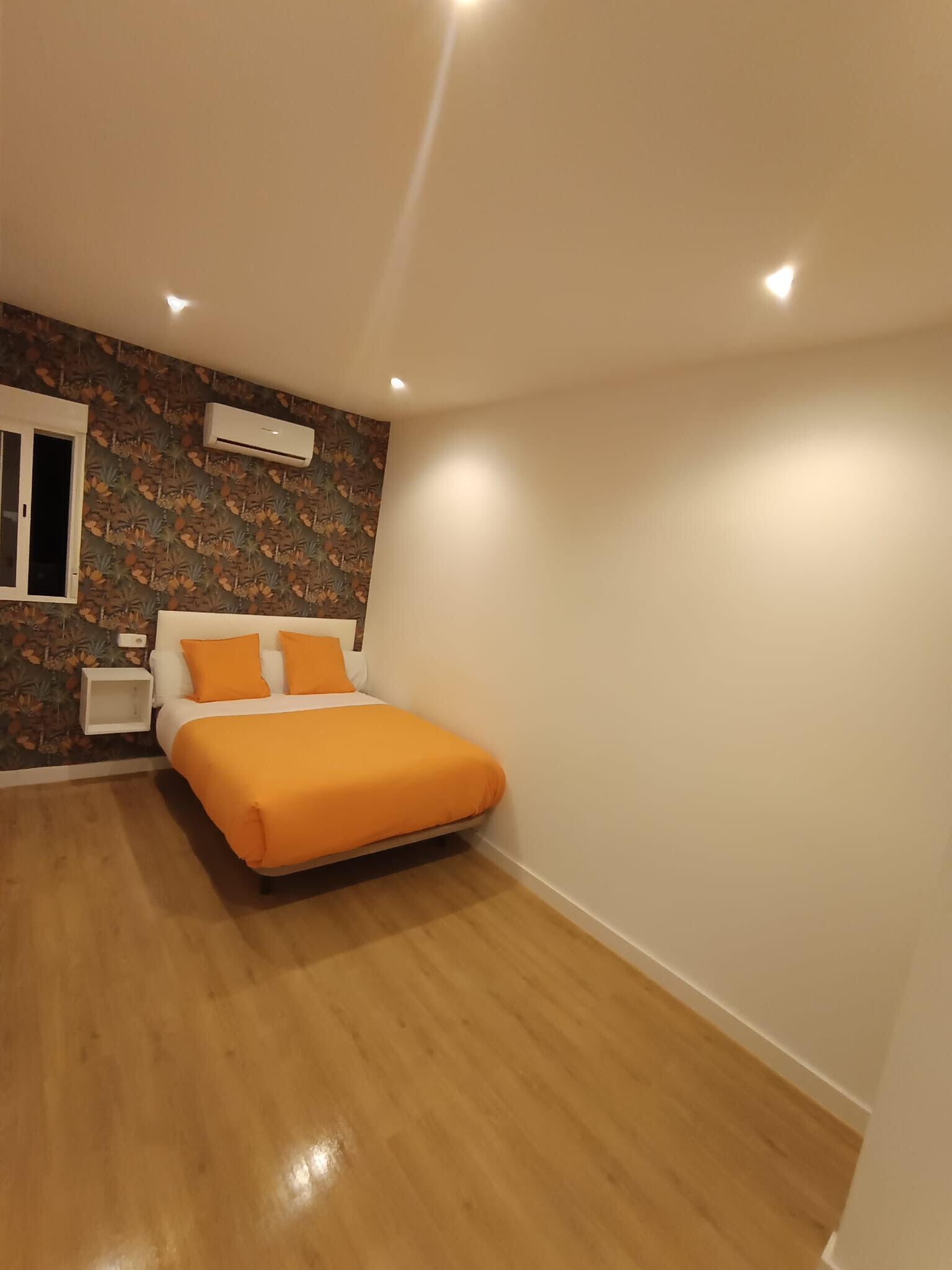 A bedroom with a bed, a nightstand, a wall-mounted air conditioner, and a window.
