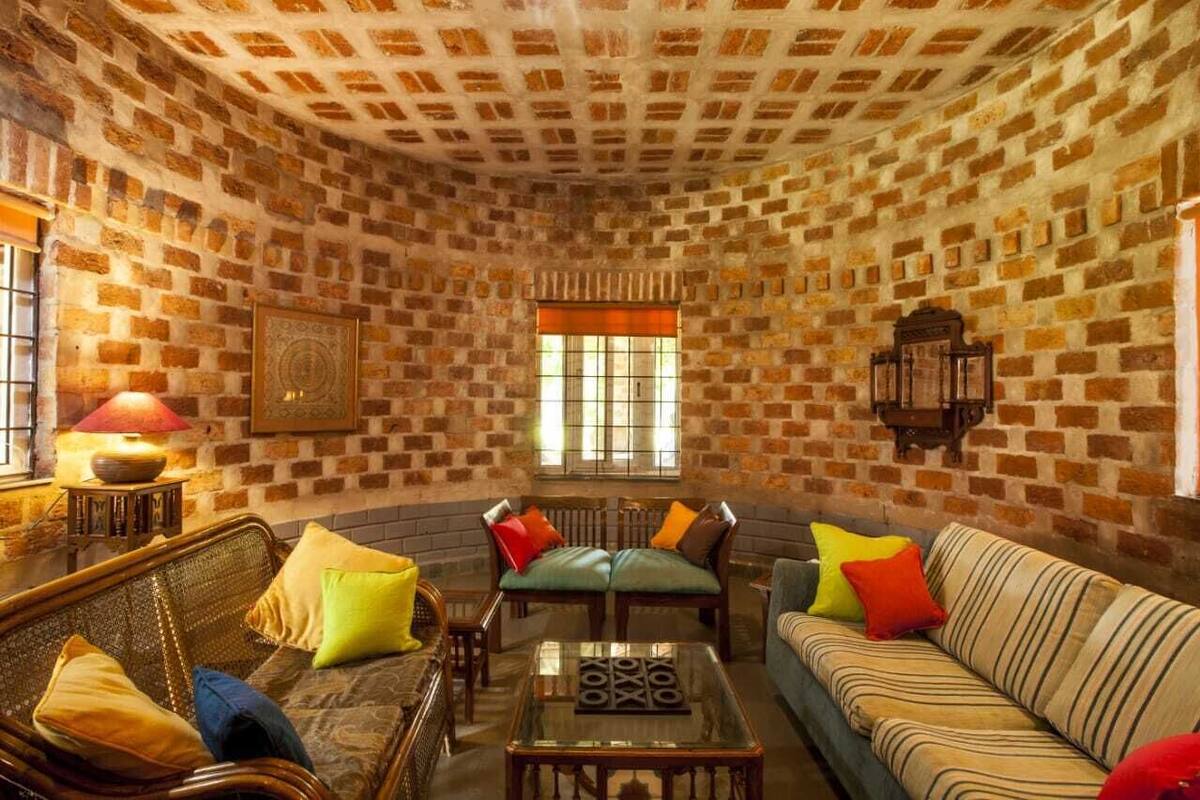 Une pièce chaleureuse avec des murs en briques, un plafond en bois et une grande fenêtre dotée de stores orange. On y trouve deux canapés, une petite table et un fauteuil.