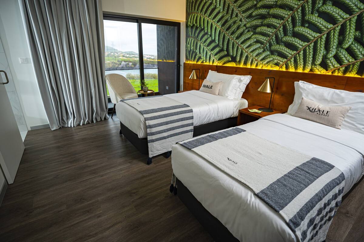A hotel room with two beds, a large window with a view of the outdoors, a headboard with a leaf pattern, and a nightstand with a lamp.