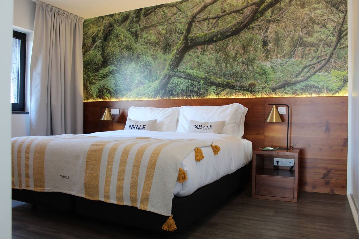 A bedroom with a bed, a nightstand, a wall-mounted lamp, and a window with curtains.