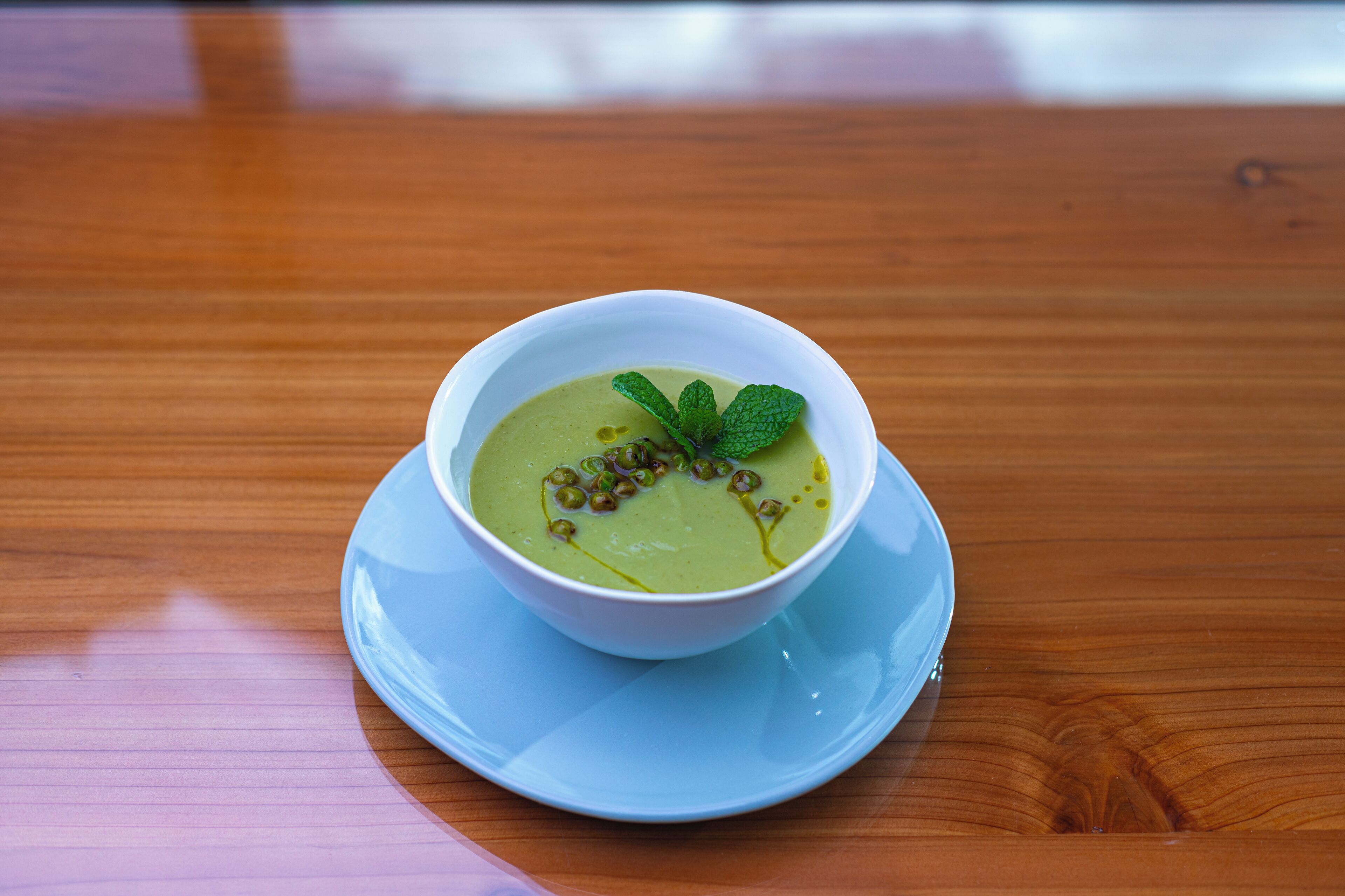 A bowl of green soup on a white plate with a mint garnish, placed on a blue saucer on a wooden table.