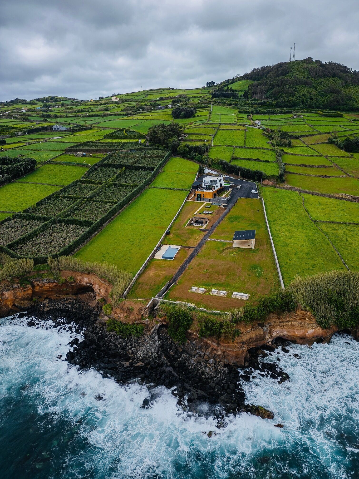 An aerial view of a coastal area with terraced fields, a small building with solar panels, and a rocky coastline with waves crashing against the shore.