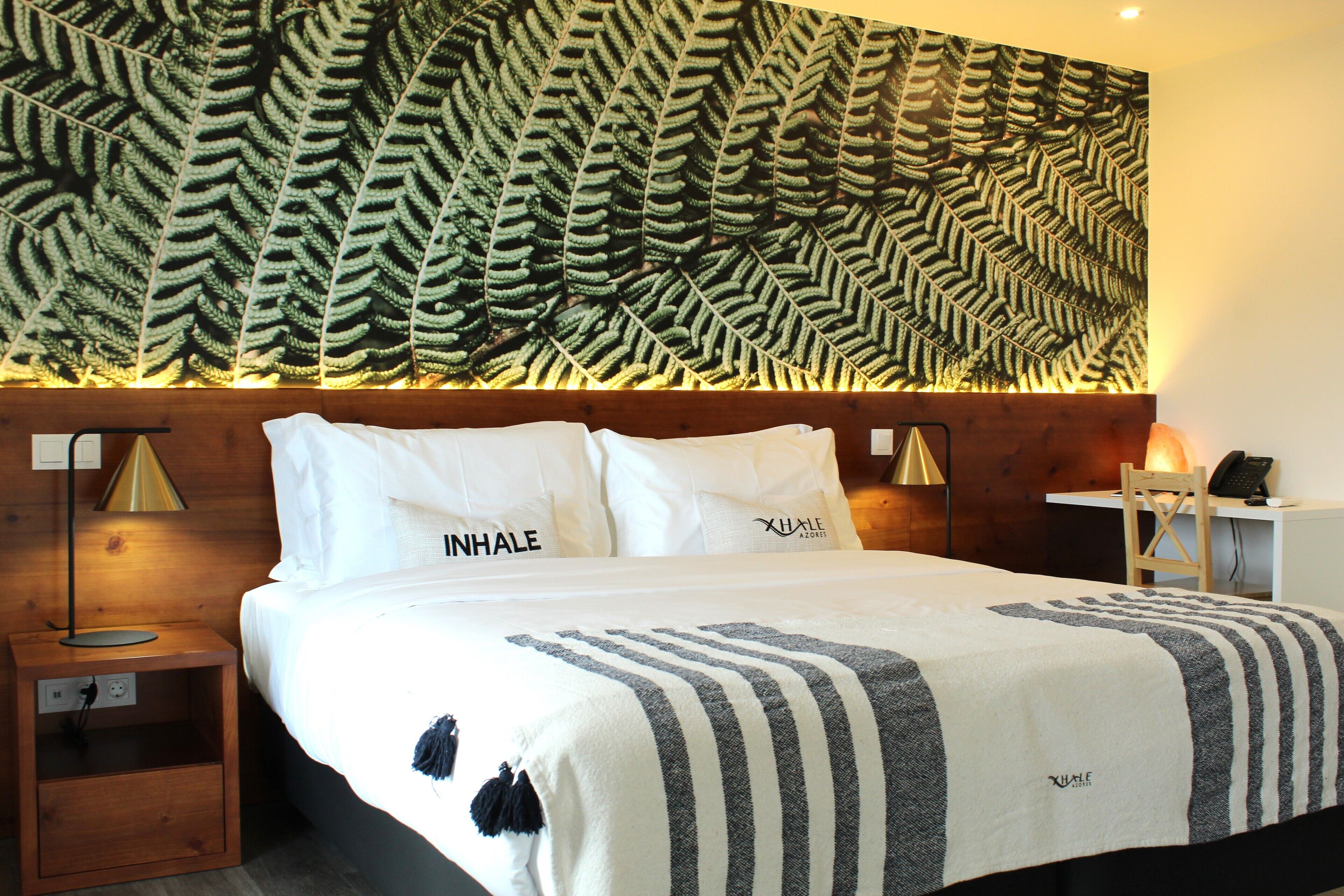 Una habitación de hotel con cama, mesita de noche, escritorio y un gran papel tapiz con estampado de hojas de helecho.
