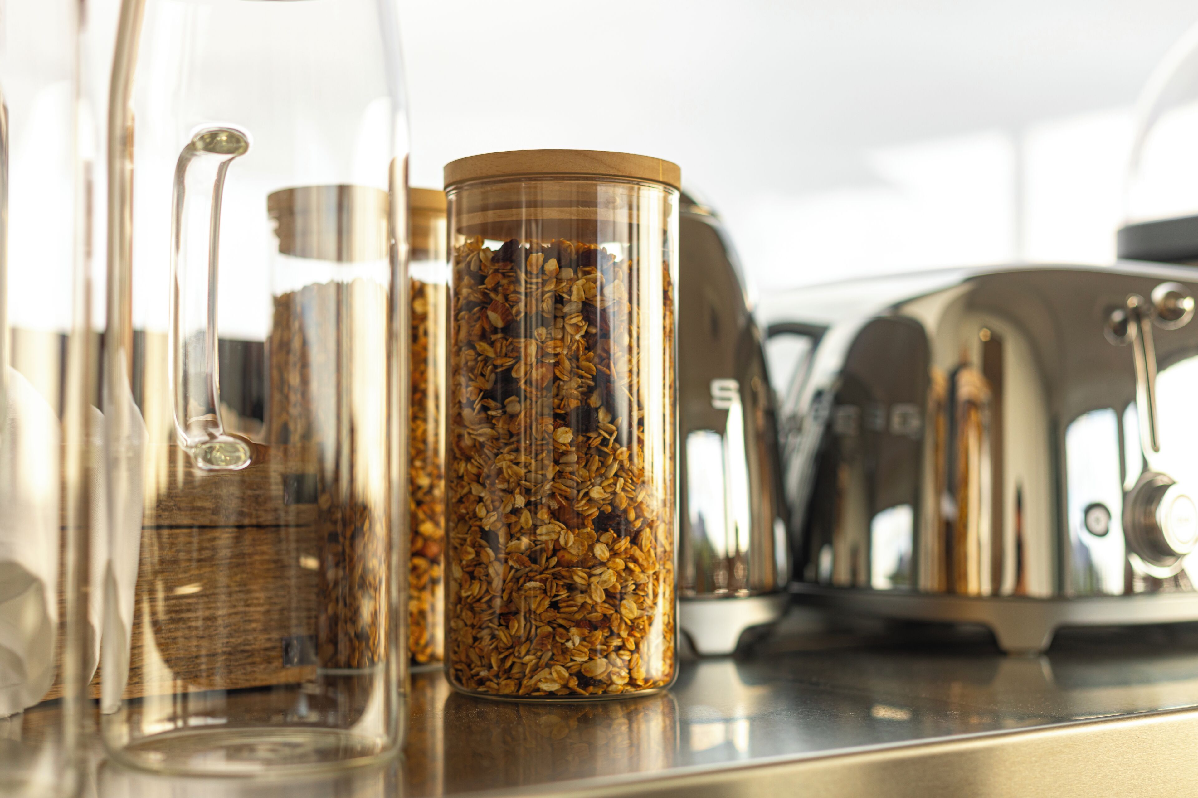 Una encimera de cocina con varios recipientes de vidrio que contienen granos y semillas, un hervidor de acero inoxidable y una jarra de vidrio.