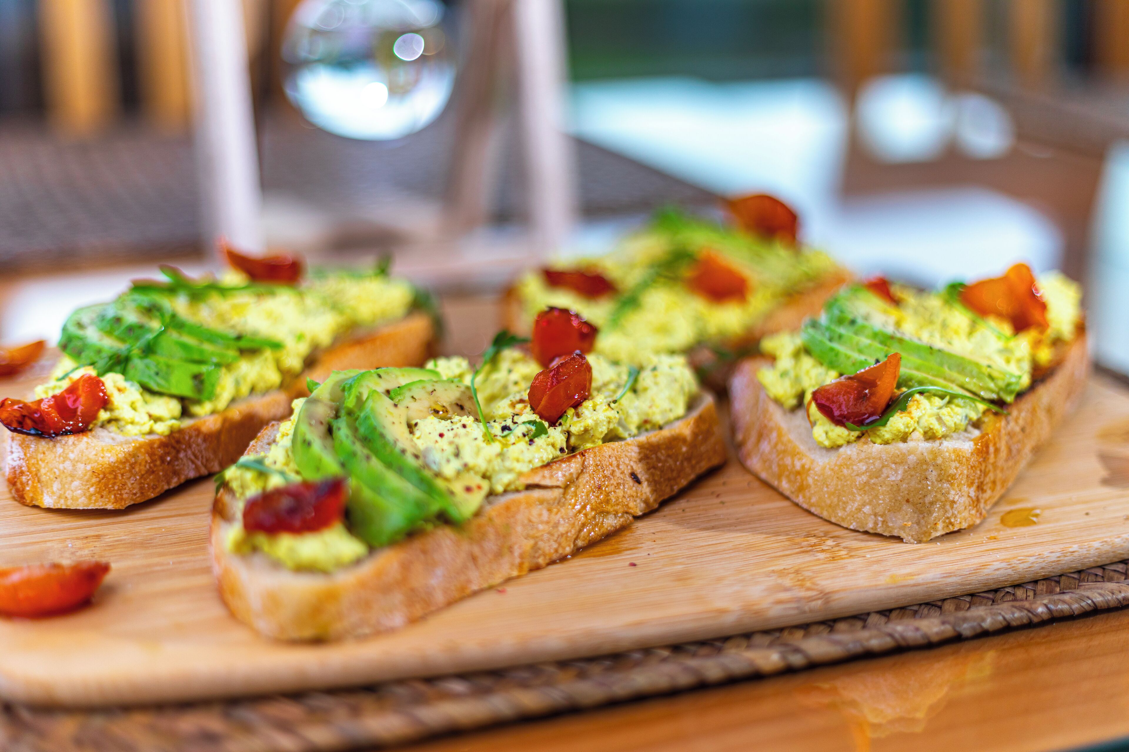 A wooden cutting board with several pieces of toast topped with avocado, bacon, and tomato slices.