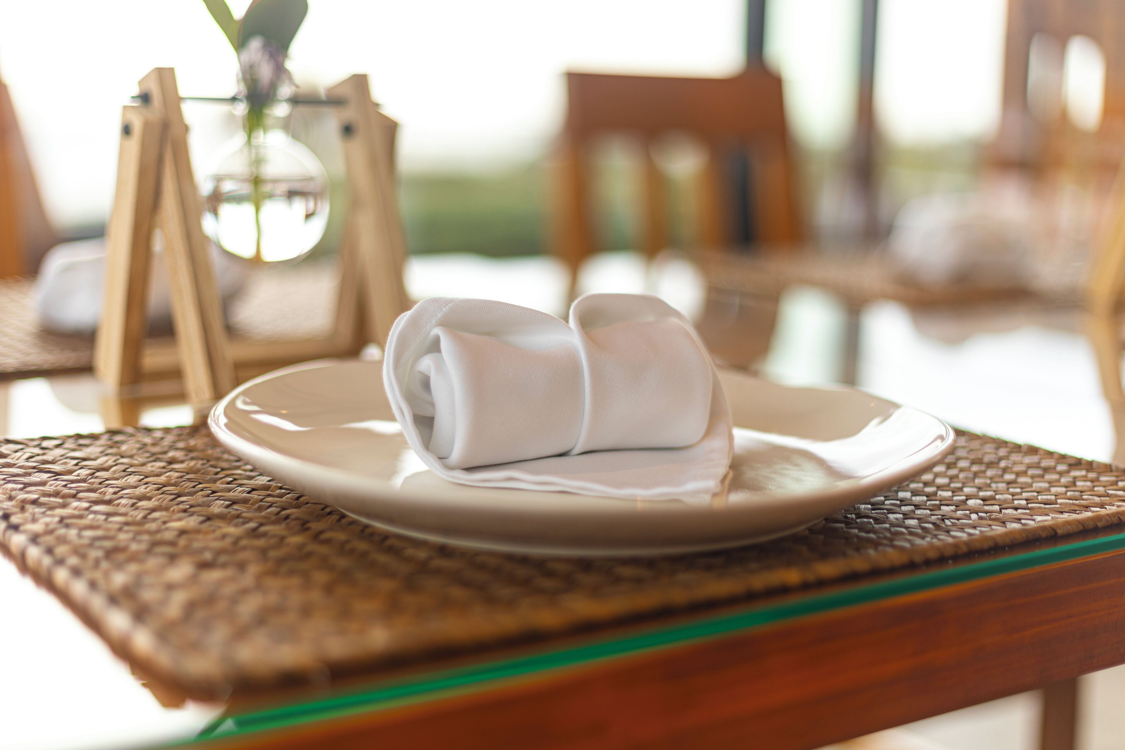 Una mesa de comedor con un plato de servilletas dobladas, un posavasos tejido y una jarra de cristal con flores.