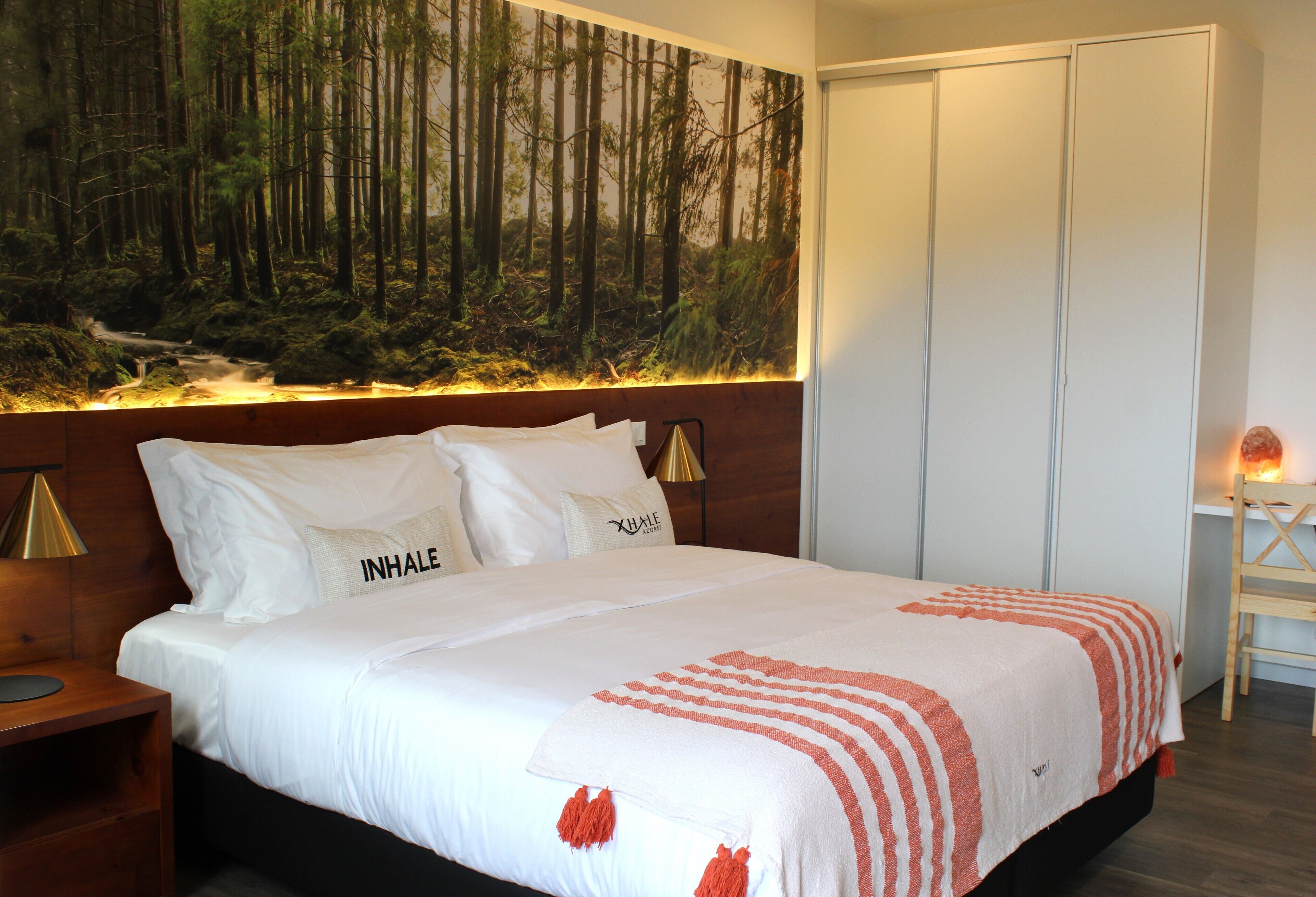 A neatly made bed with white linens and a red and white striped blanket, a wall-mounted painting of a forest, and a wooden nightstand with a lamp.