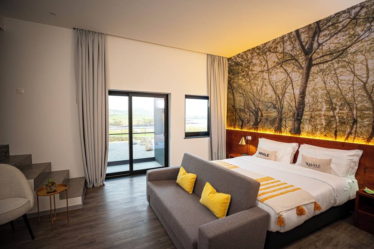 A modern hotel room with a large bed, a sofa, a small table, and a view of the outdoors through a sliding door.