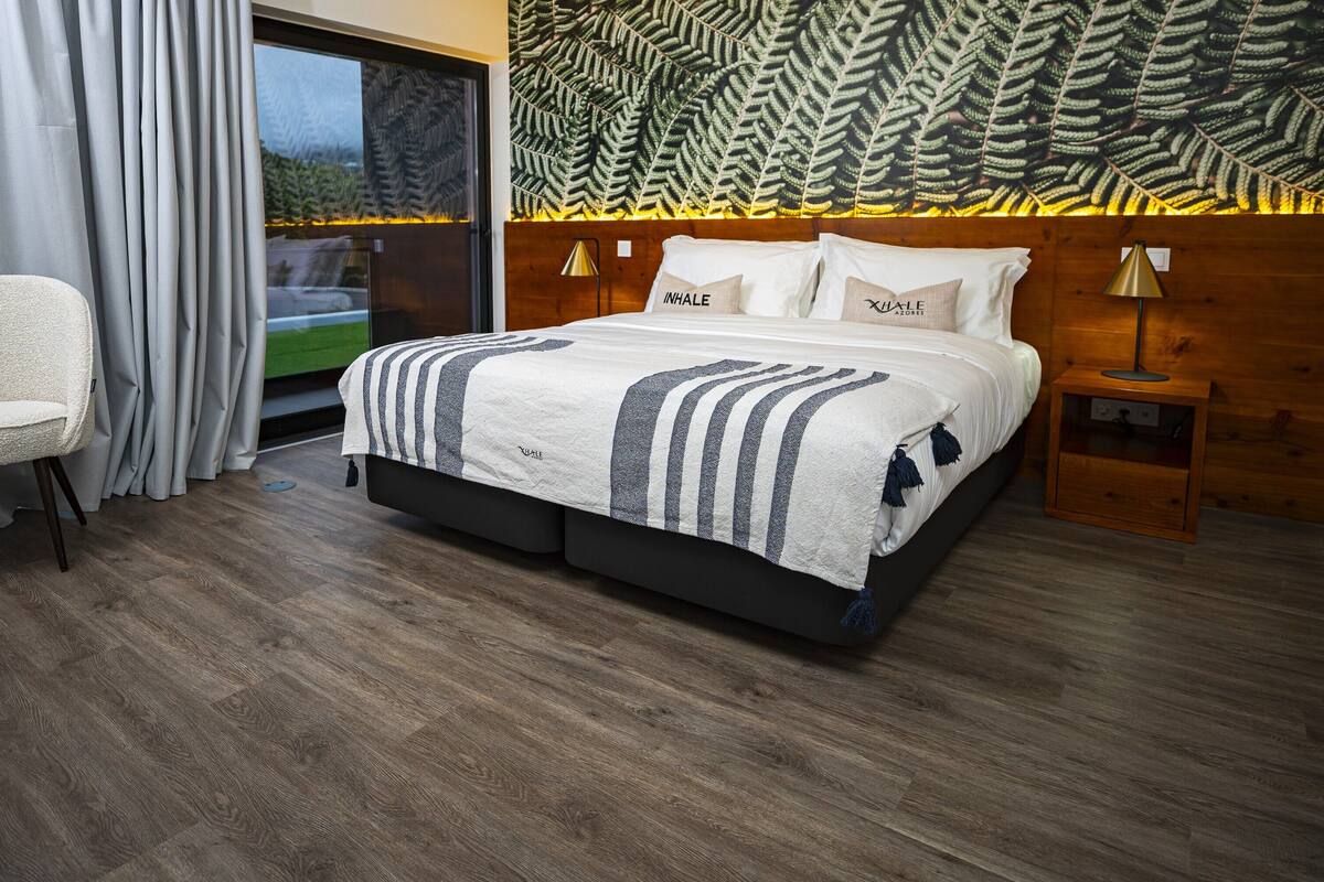 A hotel room with a bed, a chair, a nightstand, and a large window with curtains.