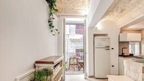 A narrow hallway leading to a balcony with a view of a red building. A white refrigerator and a wooden table are visible in the room.