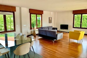 A modern living room with a glass dining table, a grey sofa, a yellow armchair, and a TV. - 1A119 El Sangar Somio (Gijón)