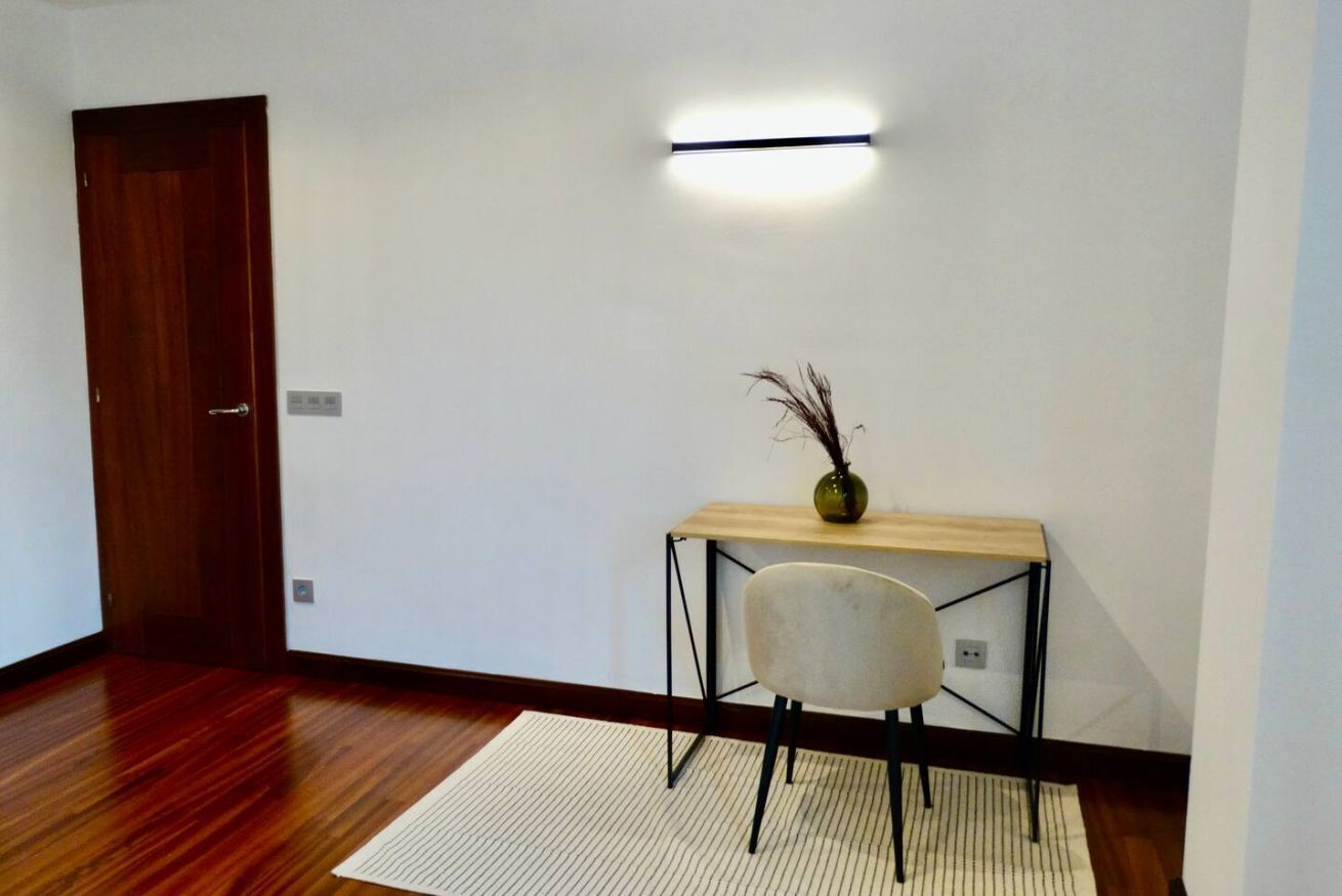 A room with a wooden floor, a white wall, a wooden door, a desk with a chair, a vase with dried plants, and a wall-mounted light fixture.