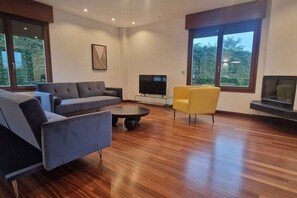 Een moderne woonkamer met een bank, fauteuils, een salontafel, een televisie en grote ramen met uitzicht op groen.