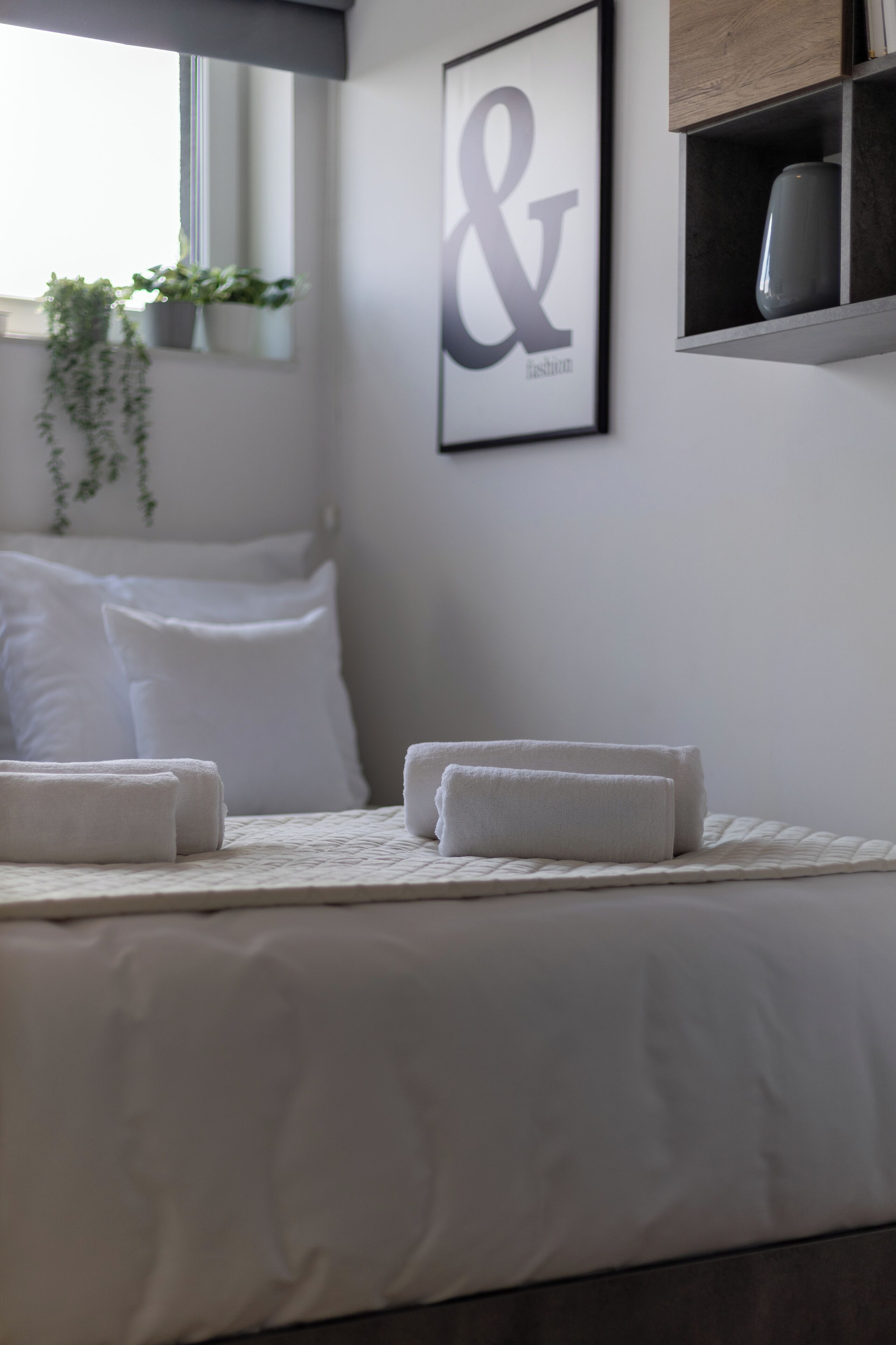 A neatly made bed with white linens and two folded towels, a framed sign with "& fashion" on the wall, and a hanging plant near a window.