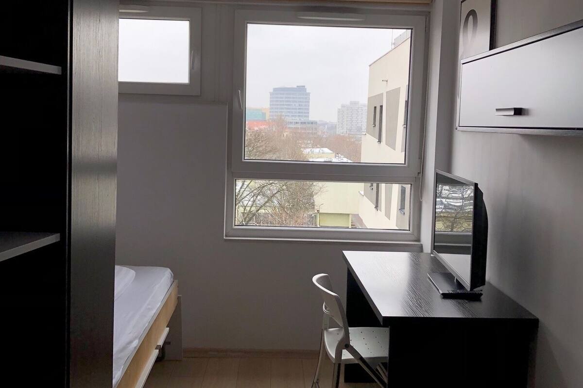A small, modern room with a single bed, a desk, and a chair. A large window provides a view of buildings outside.