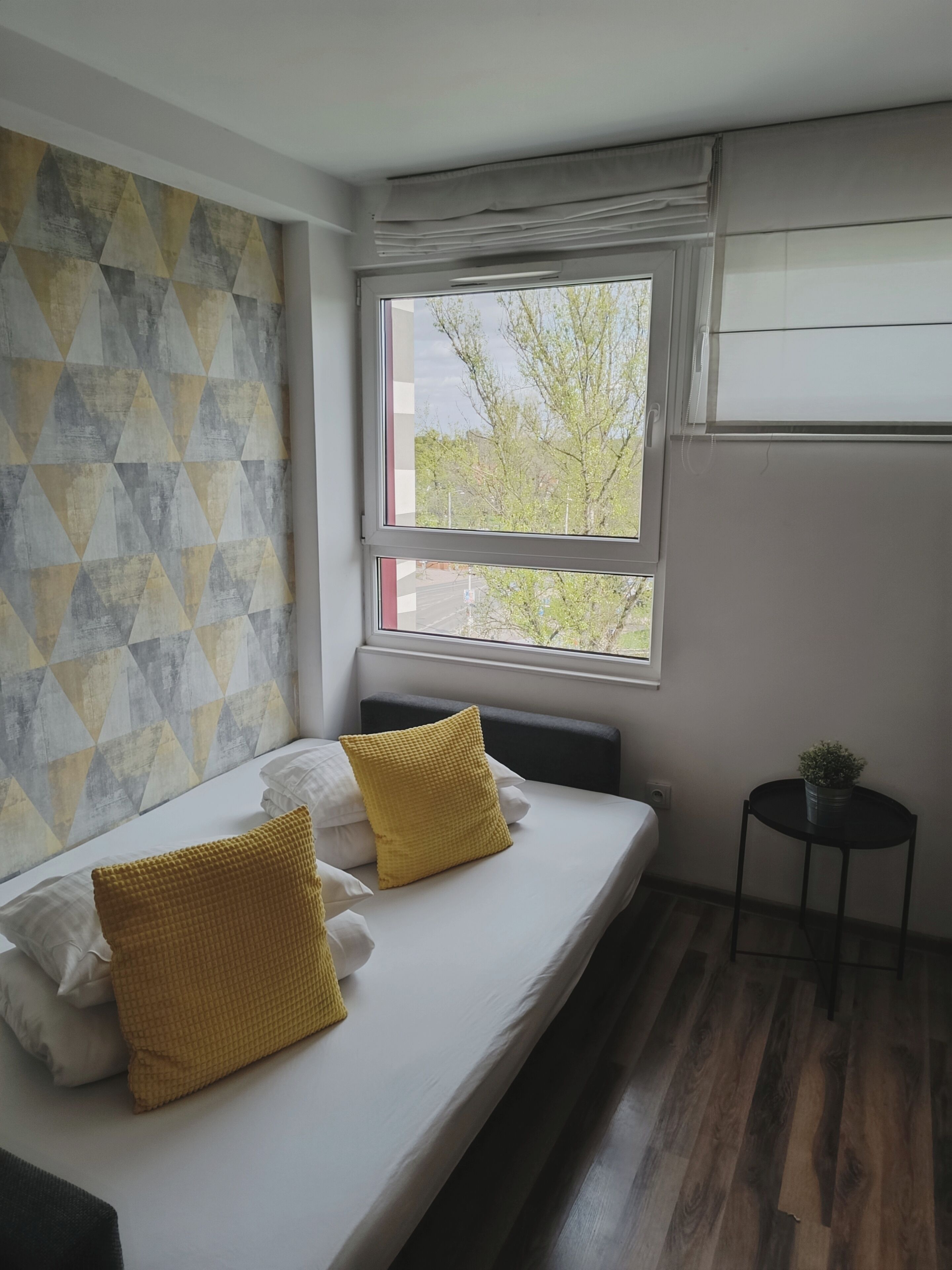 A bedroom with a bed, pillows, a window, a small table, and a wall with a geometric pattern.