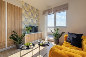 En moderne stue med en gul sofa, et fjernsyn og et spiseområde med en bakke æbler og glas.