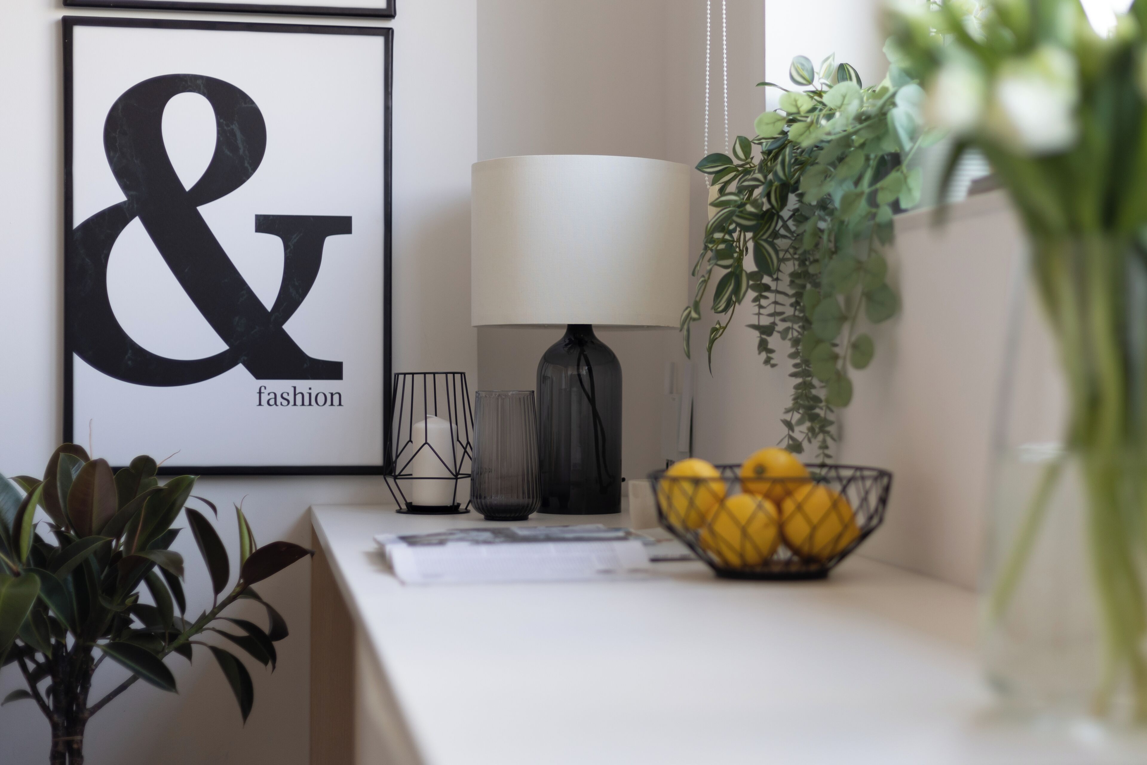 A neatly arranged room with a framed sign, a lamp, a vase with green plants, and a bowl of lemons on a table.