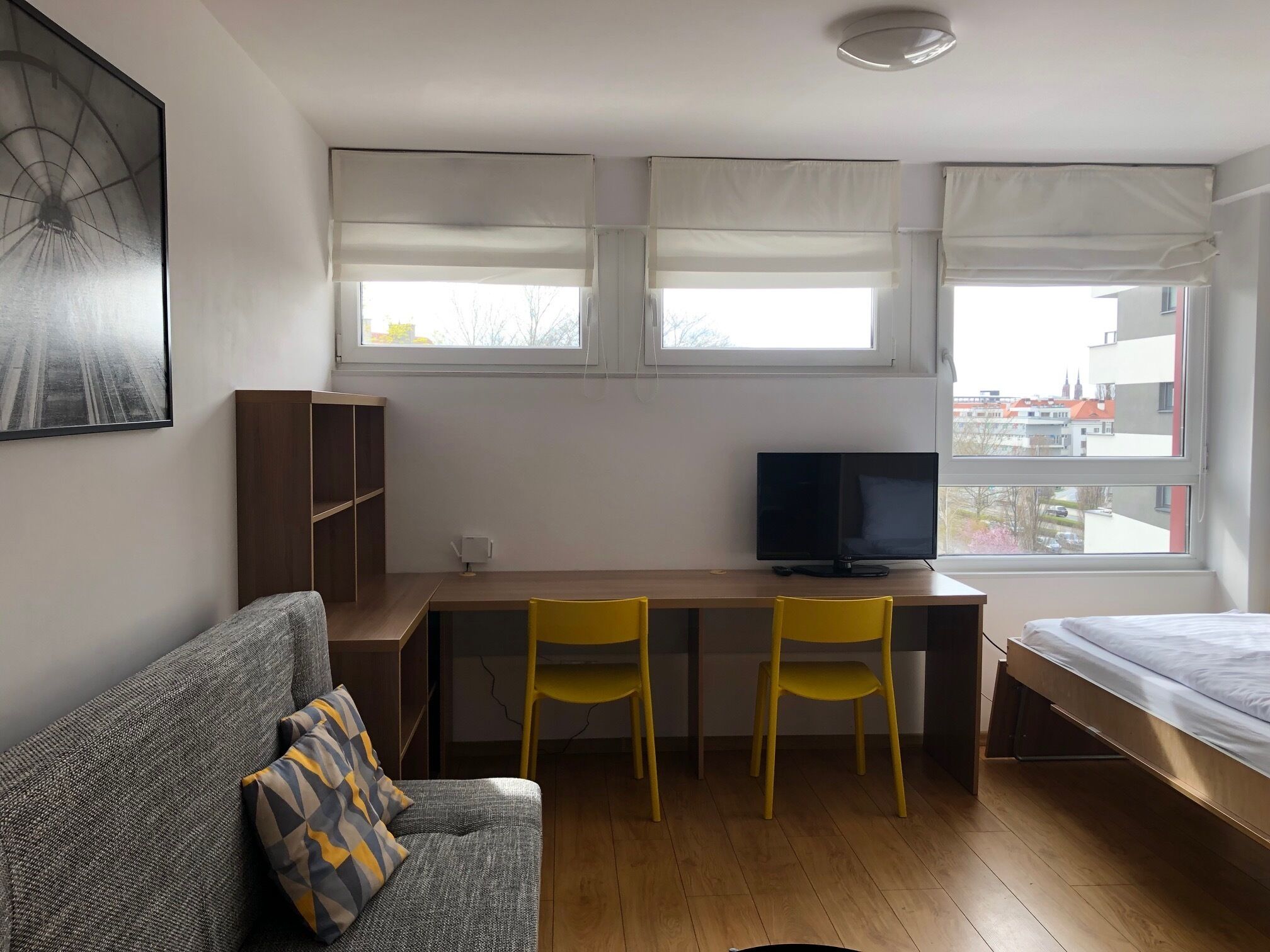 Ein gemütlicher Raum mit einem Bett, einem Schreibtisch mit Fernseher, zwei gelben Stühlen, einem grauen Sofa und einem Bücherregal.