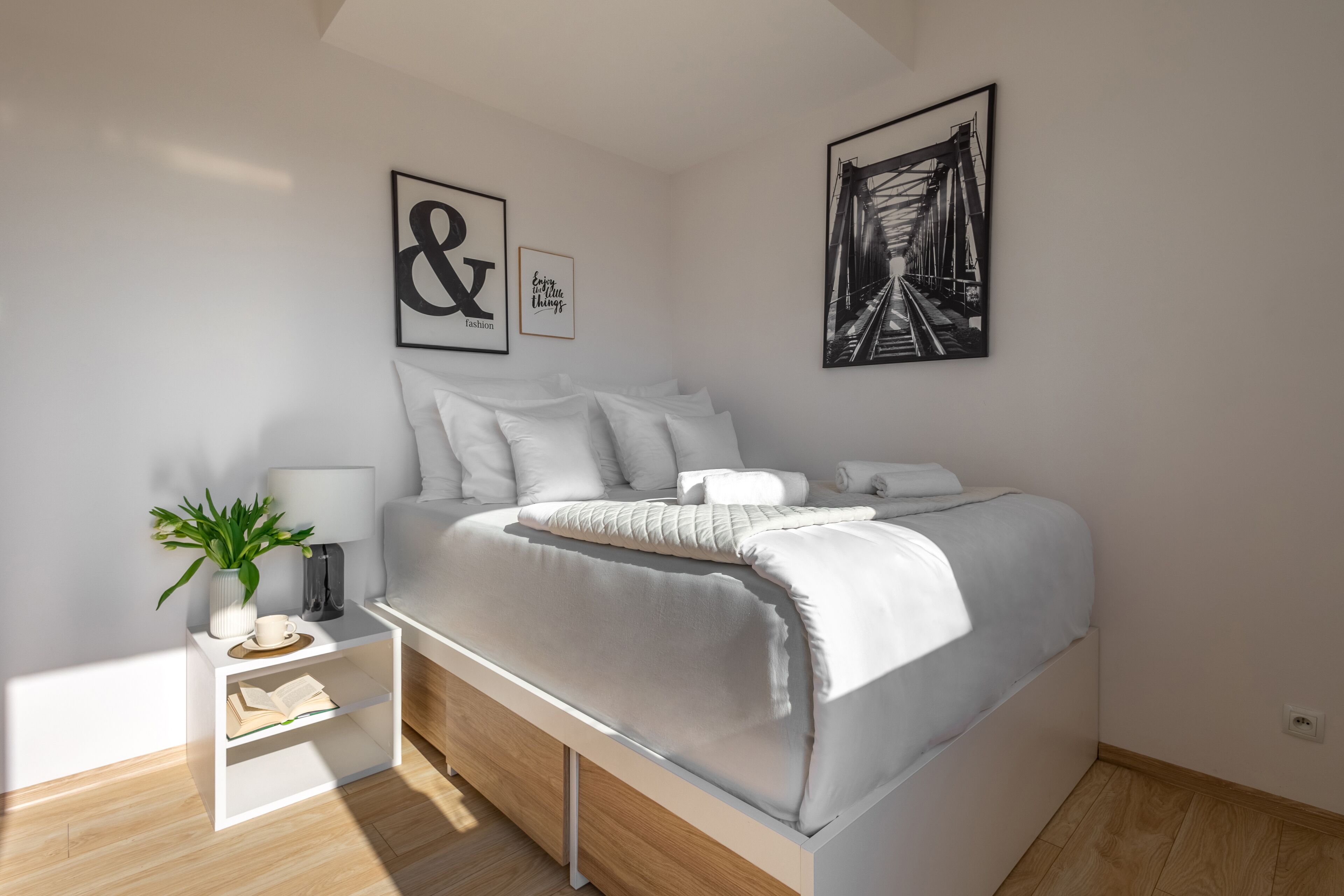 A modern bedroom with a bed, bedside tables, a nightstand, a potted plant, and artwork on the walls.