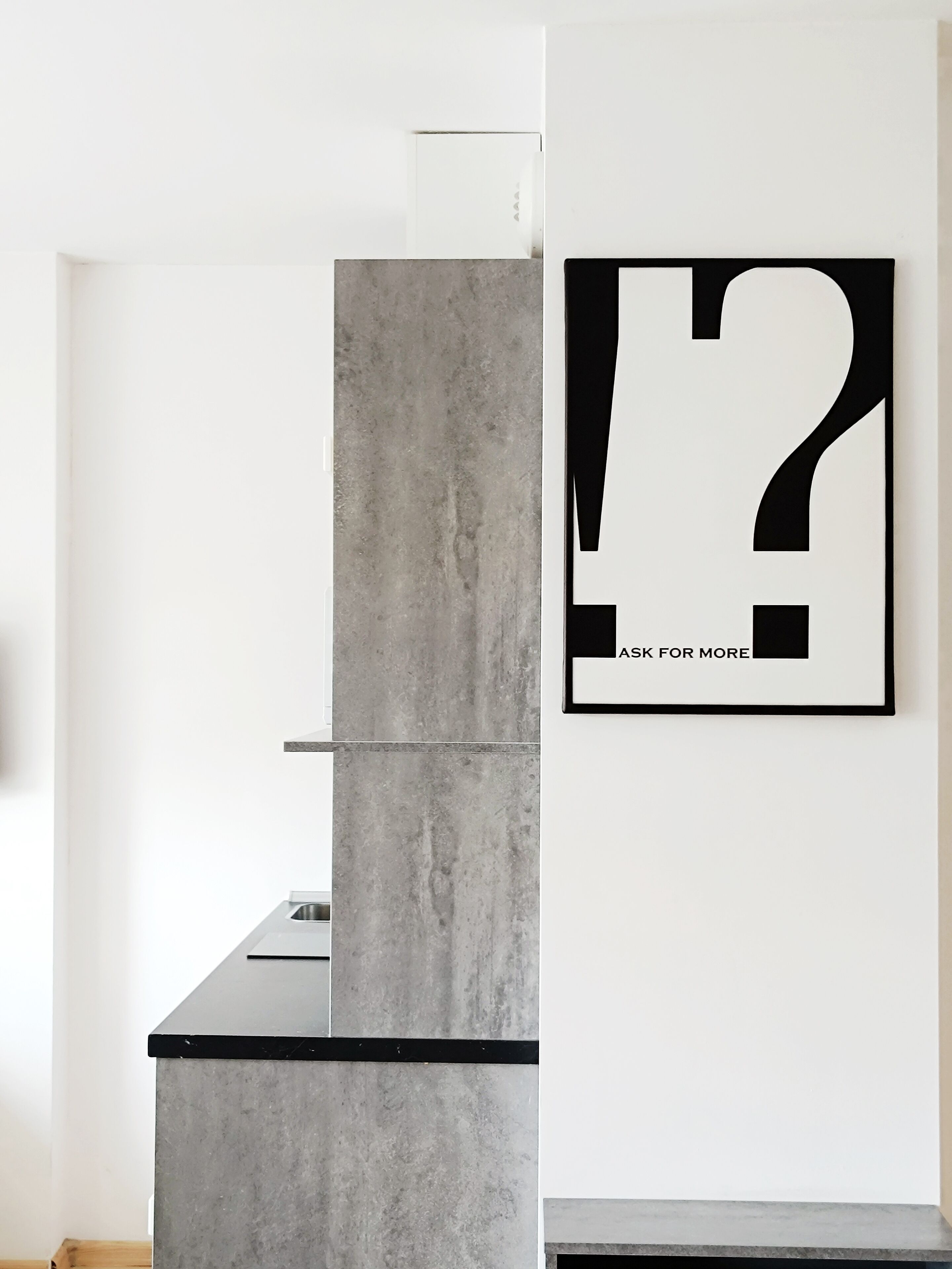 Ein modernes Interieur mit einer grauen Betonstütze, einer schwarzen Arbeitsplatte und einem gerahmten Fragezeichen an der Wand.