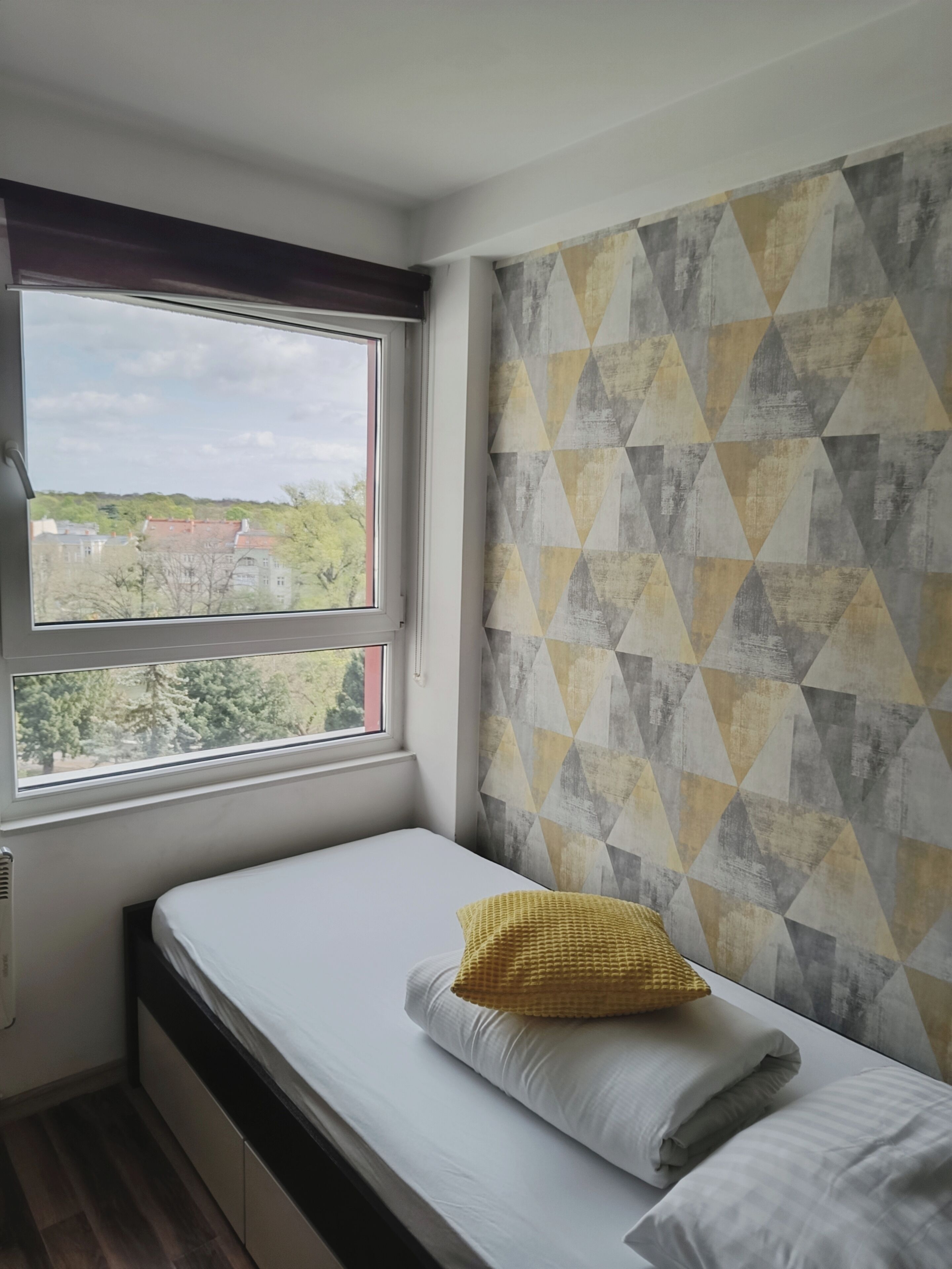 Una habitación pequeña con una cama individual, una ventana que ofrece vistas a edificios y árboles, y una pared con un diseño geométrico.