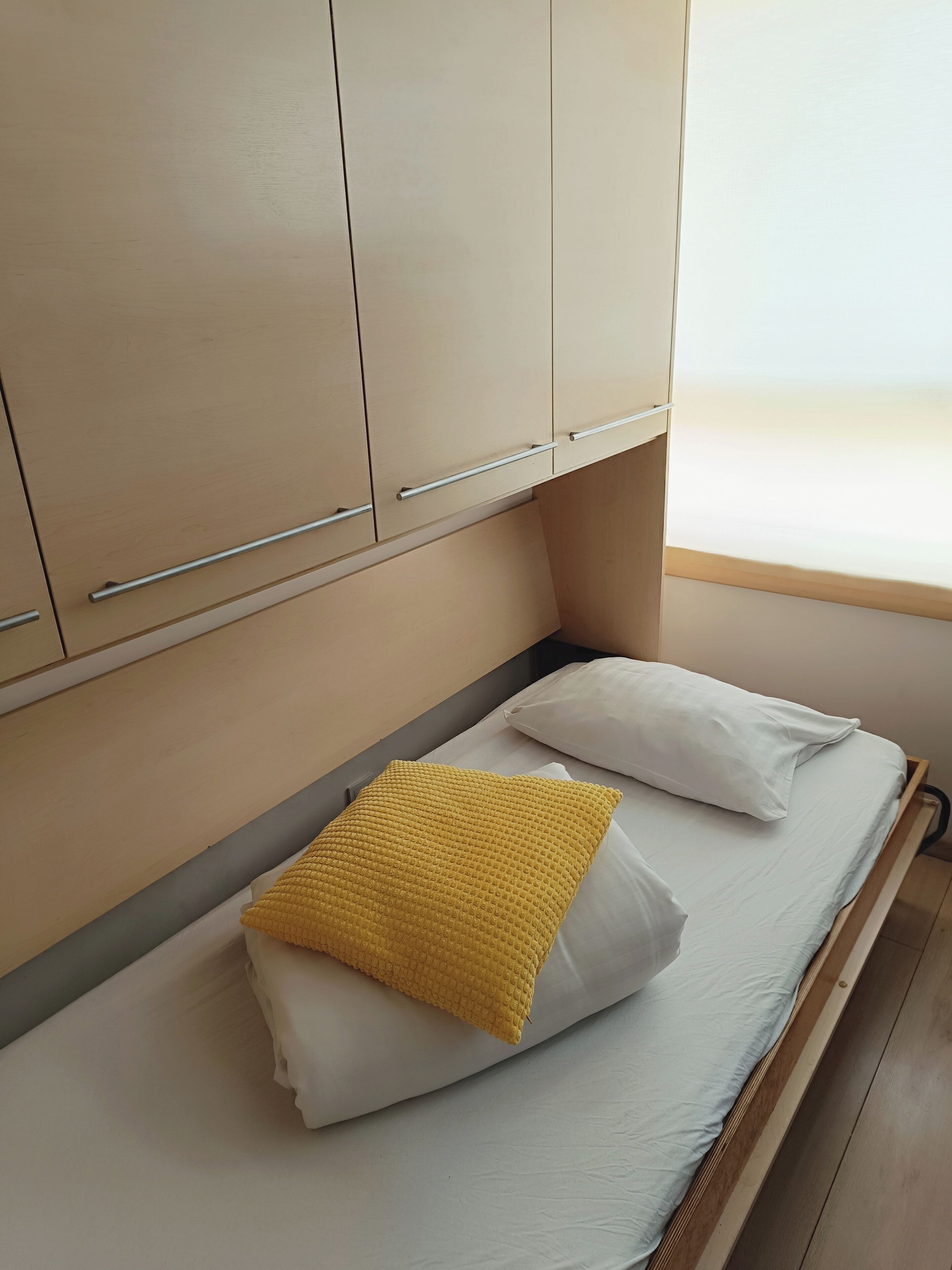 A single bed with white bedding and a yellow textured pillow, a wooden headboard, and beige cabinets with silver handles.