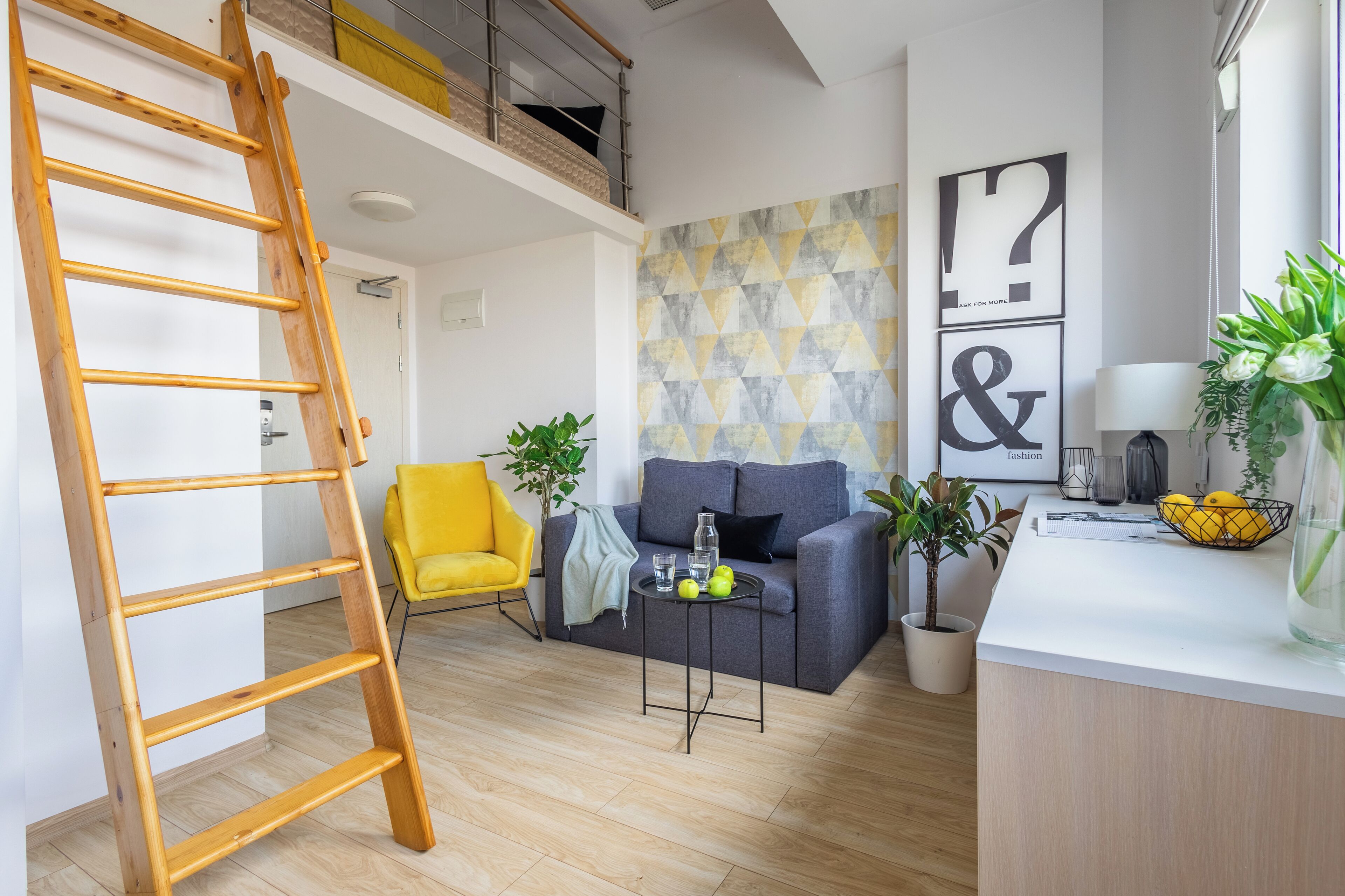 A modern living room with a wooden ladder, a yellow chair, a grey sofa, and a small table with apples and a bottle.
