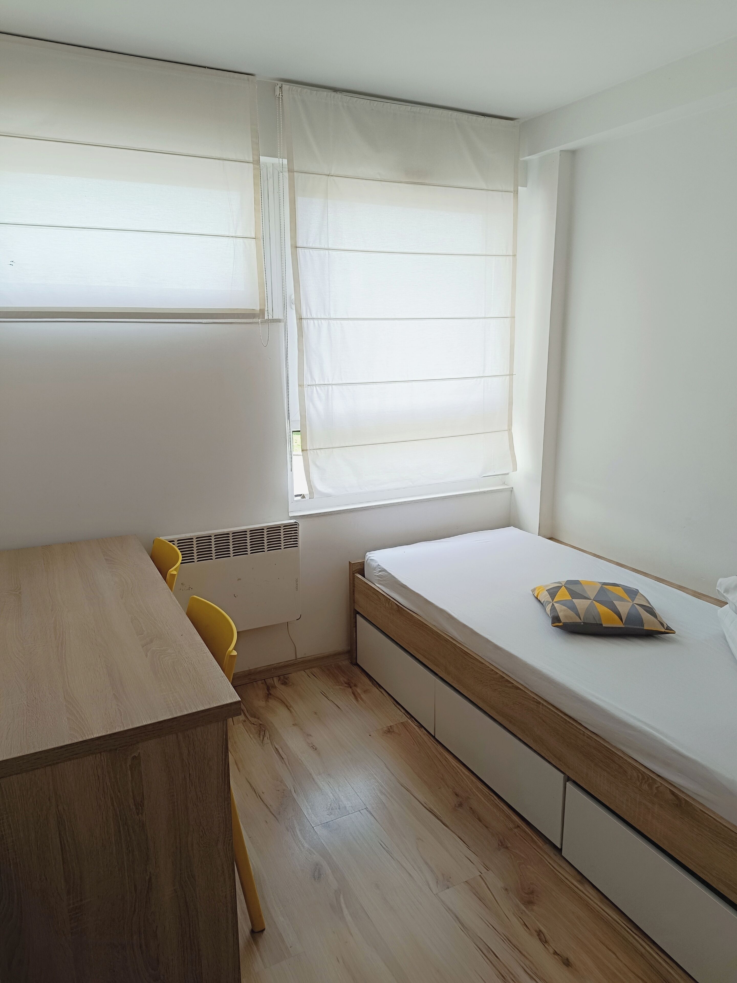 A small, well-lit room with a wooden table, yellow chairs, a bed with a geometric pillow, and a window with white curtains.