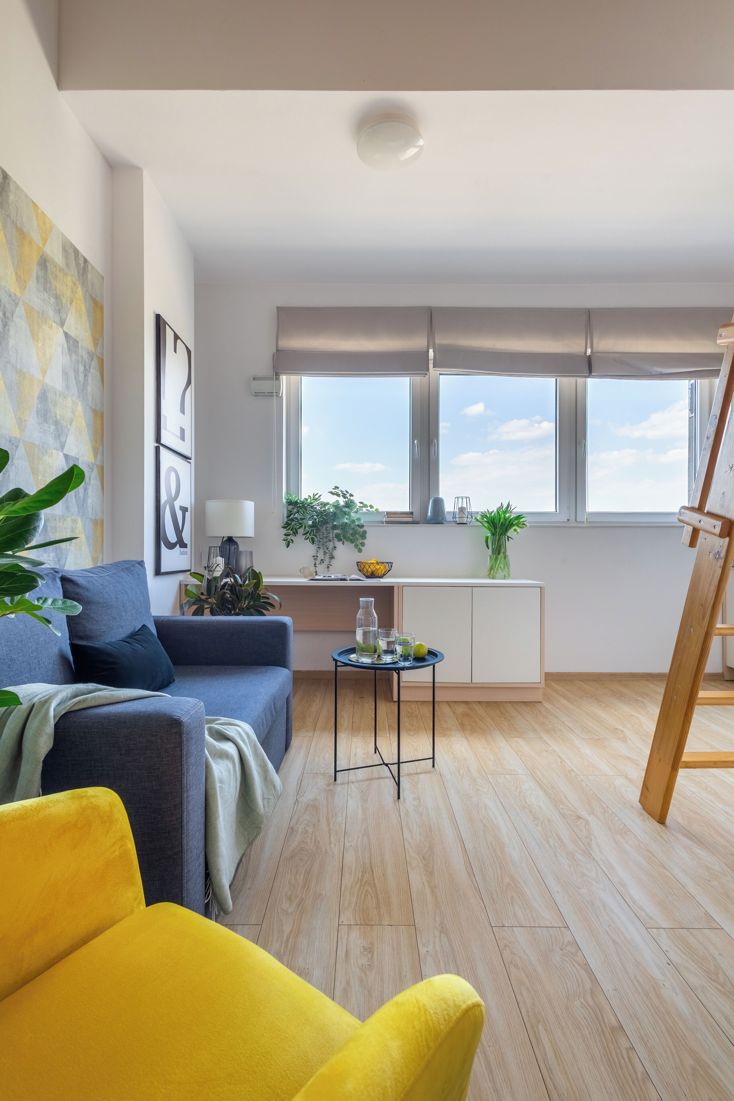 A modern living room with a sofa, a yellow armchair, a small table with drinks, and a painting on the wall.
