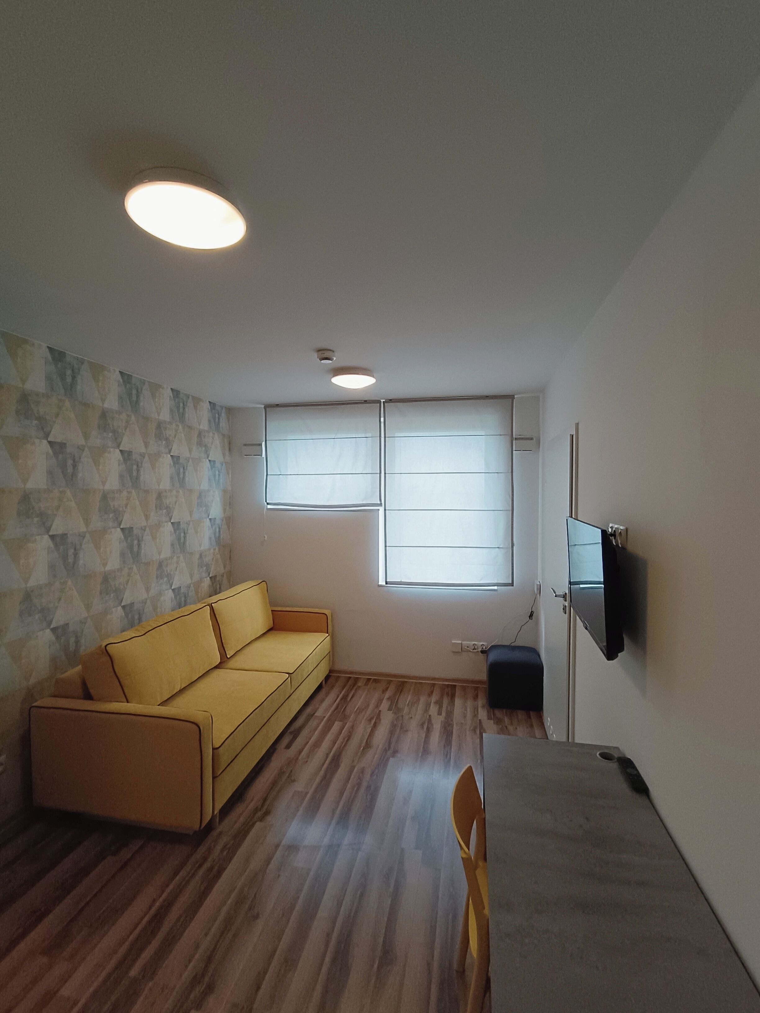 A small living room with a yellow sofa, a flat-screen TV mounted on the wall, and a window with blinds.