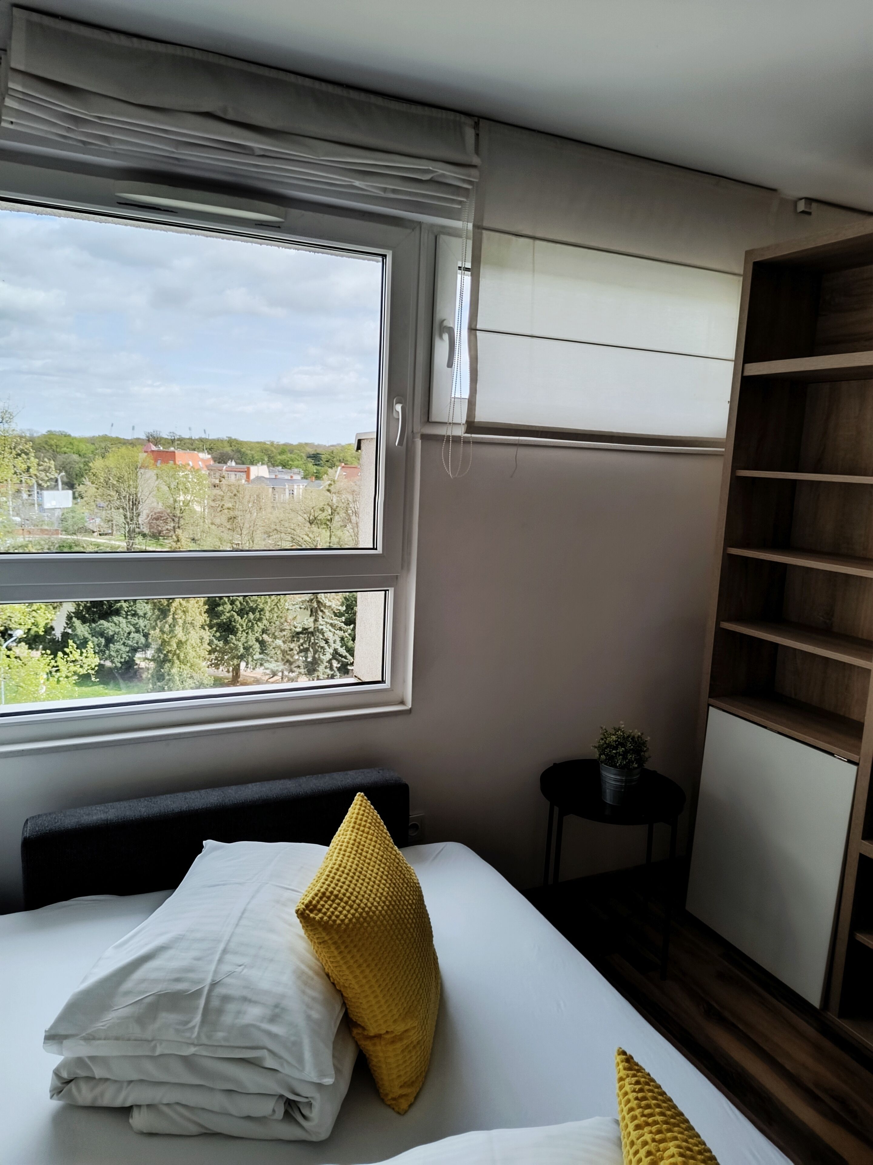 Ein Schlafzimmer mit großem Fenster, einem Bett mit weißer Bettwäsche und einem gelben Kissen, einem kleinen Tisch mit Pflanze und einem Holzschrank.