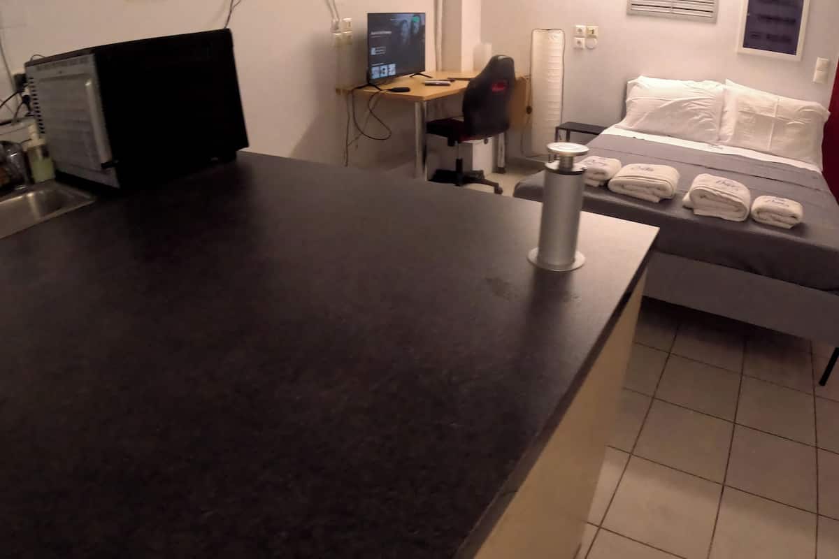 A small hotel room with a kitchenette, a desk with a computer, a chair, a bed with white linens, and a red door.