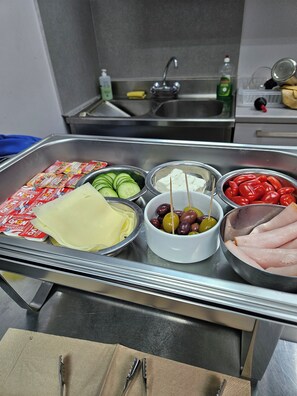 A stainless steel buffet table with various food items including cheese, olives, cucumbers, and sliced meat.