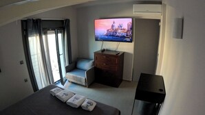 A hotel room with a bed, a TV on a wooden cabinet, a chair, and a window with curtains. - Data Maisonettes  (Chios)
