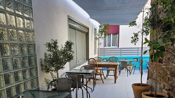 A covered outdoor patio with a glass table and chairs, potted plants, and a view of a building with a blue wall and a red awning. - Data Maisonettes  (Chios)