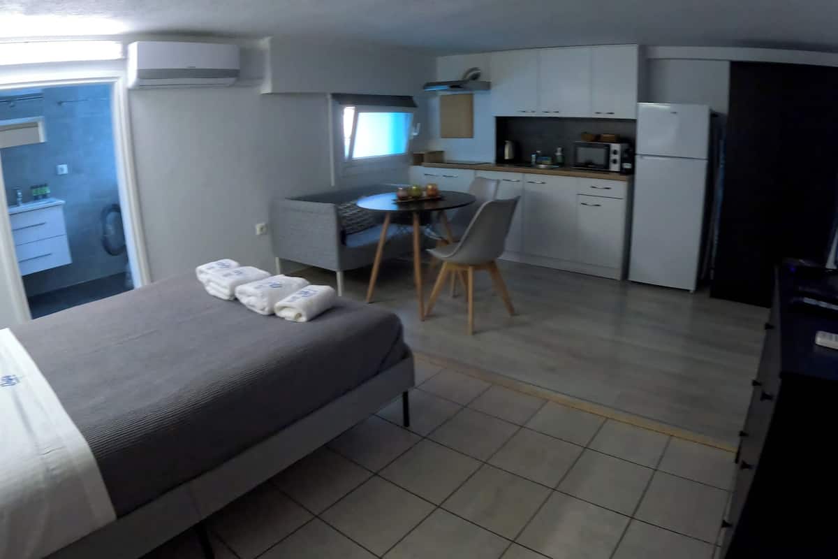 A compact hotel room with a bed, a small dining table with chairs, a kitchenette, and a TV.
