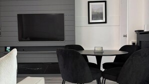 Un salon moderne avec un téléviseur gris fixé au mur, une table de salle à manger ronde entourée de chaises noires et un canapé blanc.