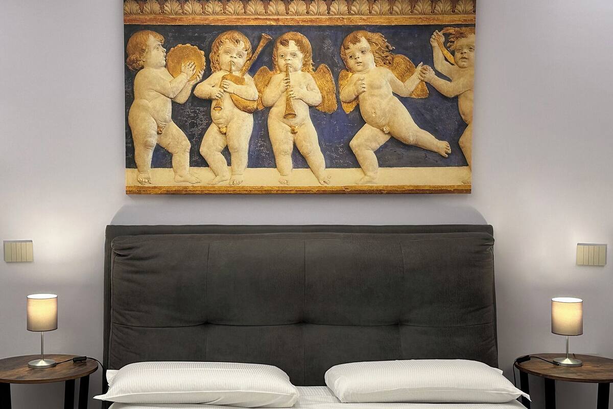 Quarto com um grande mural na parede retratando querubins tocando instrumentos musicais, uma cabeceira estofada escura, uma cama com roupa de cama branca e duas mesas de cabeceira com abajures.