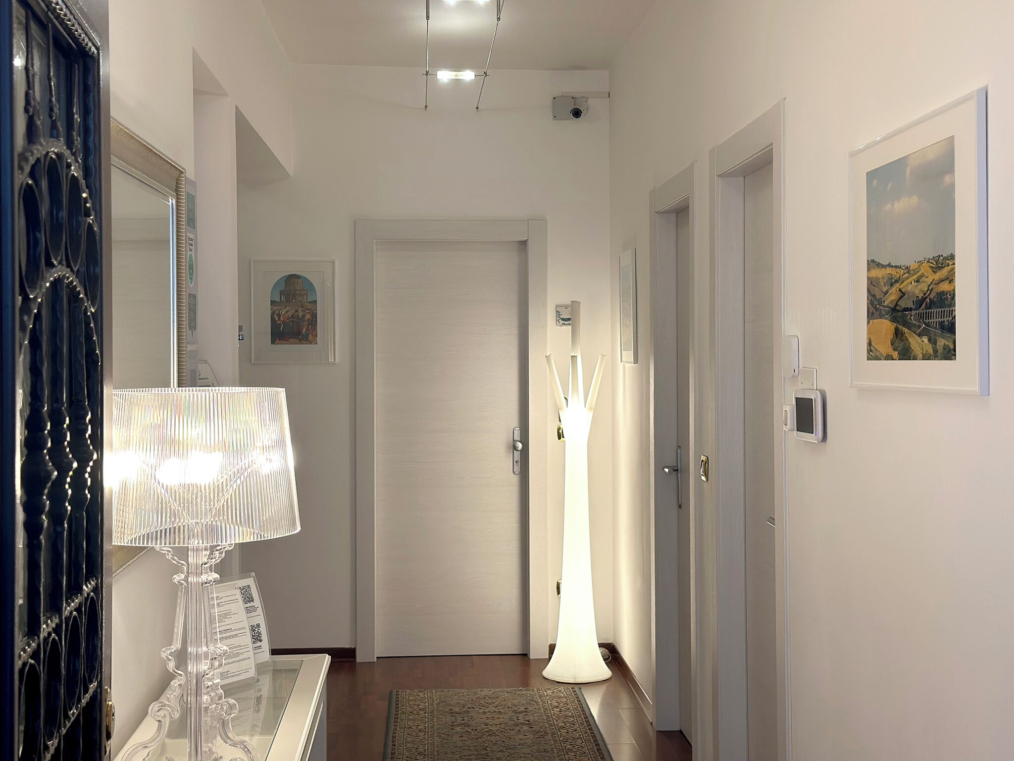 A hallway with a door, a lamp, a security camera, and a framed picture on the wall.