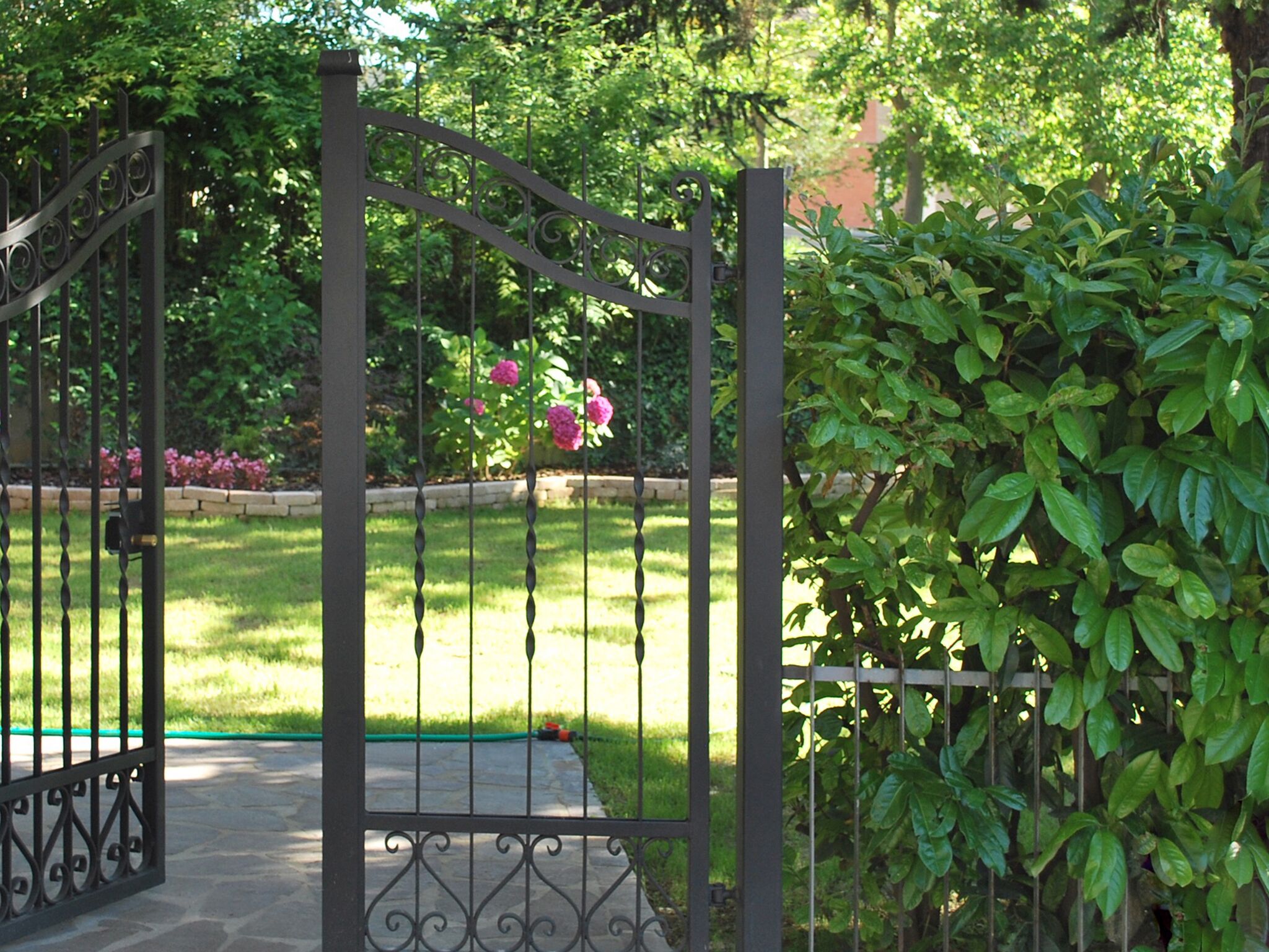 Una reja de hierro forjado con diseño decorativo, que da acceso a un jardín bien cuidado con césped y plantas en flor.